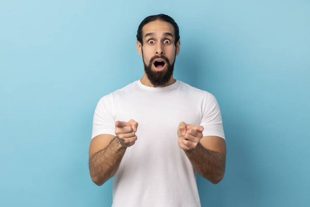 Omg just look there. Portrait of shocked astonished man with beard wearing white T-shirt pointing directly at camera cannot believe own eyes. Indoor studio shot isolated on blue background. - Photo, Image