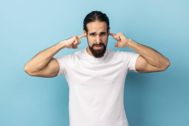 I don't want to here this. Portrait of man with beard wearing white T-shirt standing and holding fingers on his ear, unpleasant sounds. Indoor studio shot isolated on blue background. - Fotó, kép