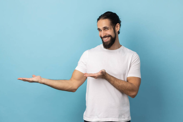 Portrait of satisfied optimistic handsome man with beard in white T-shirt standing with wide raised arms and welcoming or sharing. Indoor studio shot isolated on blue background. - Fotó, kép
