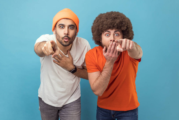 Wow, look at this. Portrait of two astonished surprised young adult hipster men looking at camera with big eyes, pointing at camera with fingers. Indoor studio shot isolated on blue background. - Φωτογραφία, εικόνα