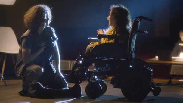 An actor kneeling next to a disabled actress in a wheelchair acting a romantic scene of a play in the theater, during a rehearsal with the director - Photo, Image