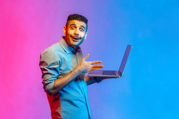 Portrait of amazed handsome man in shirt watching shocked video, having astonished facial expression, looking at camera with big eyes. Indoor studio shot isolated on colorful neon light background. - Photo, image