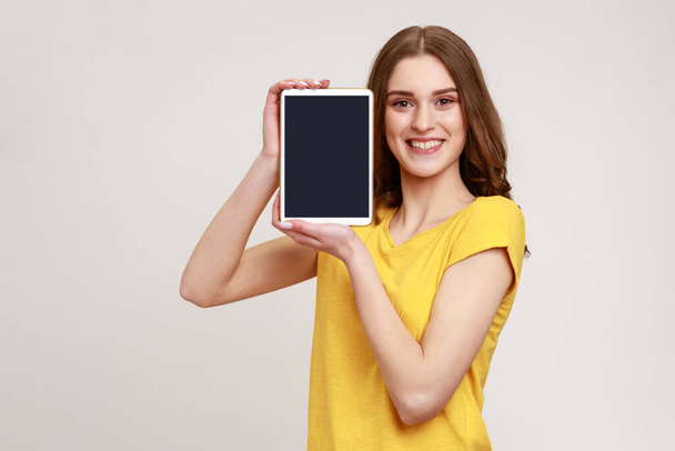 Portrait of beautiful smiling young teenage with brown hair standing and holding tablet empty screen, sowing device and looking at camera. Indoor studio shot isolated on gray background. - Fotoğraf, Görsel
