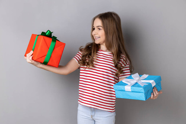 Portrait of charming little girl wearing striped T-shirt celebrating her birthday, holding two present boxes in hands, being happy with gifts. Indoor studio shot isolated on gray background. - Photo, Image