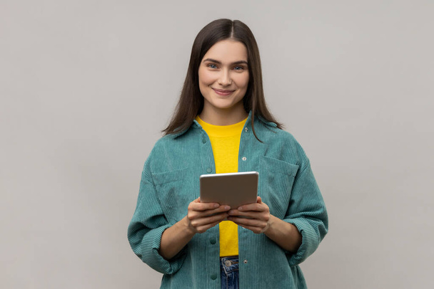 Woman with dark hair working online, using tablet for checking social networks, looking at camera with positive expression, wearing casual style jacket. Indoor studio shot isolated on gray background. - Foto, Imagen