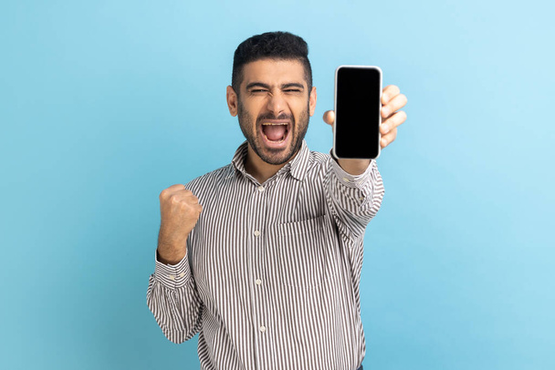 Happy businessman with beard holding smartphone and smiling making yes gesture, celebrating online lottery or giveaway victory, wearing striped shirt. Indoor studio shot isolated on blue background. - Фото, изображение