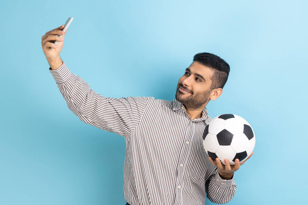 Young adult attractive businessman making selfie, having video call or broadcasting livestream with ball in hand, wearing striped shirt. Indoor studio shot isolated on blue background. - Photo, image