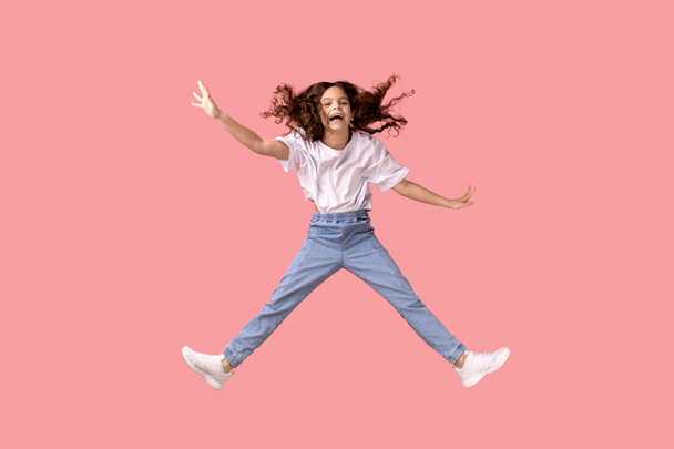 Full length portrait of excited adorable little girl wearing white T-shirt jumping in air and spread arms, smiling happily, having fun. Indoor studio shot isolated on pink background. - Foto, Bild