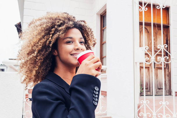 krásná elegantní afro obchodní žena pije kávu ve městě s úsměvem venku těší. kudrnaté vlasy žena pití kávy v dopoledních hodinách na sobě elegantní. - Fotografie, Obrázek