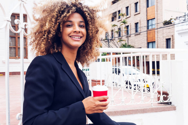 elegant business woman sitting in the city drinking coffee from her glass outdoors smiling, cheerful curly-haired afro woman drinking coffee. - Valokuva, kuva