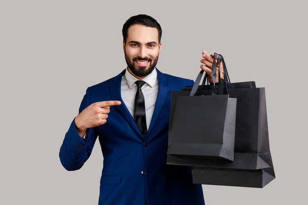 Delighted bearded man pointing finger at paper bags in hand, looking with toothy smile, shopping and sale, wearing official style suit. Indoor studio shot isolated on gray background. - Фото, зображення