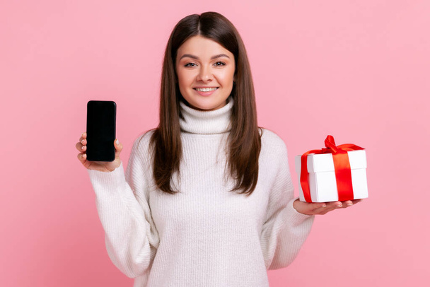 Smiling brunette woman holding wrapped present box and smart phone with blank screen for promotion, wearing white casual style sweater. Indoor studio shot isolated on pink background. - Foto, Bild