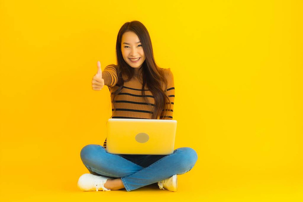Porträt schöne junge asiatische Frau sitzt auf dem Boden für den Einsatz Laptop oder Computer auf gelbem Hintergrund isoliert - Foto, Bild