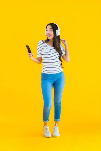 Πορτρέτο όμορφη νεαρή Ασιάτισσα γυναίκα χρησιμοποιούν έξυπνο κινητό τηλέφωνο με ακουστικά για να ακούσετε μουσική σε κίτρινο απομονωμένο φόντο - Φωτογραφία, εικόνα