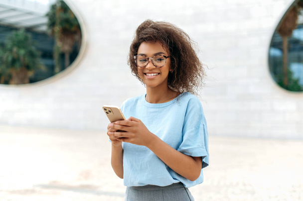 Gadget, wireless technology. Successful lovely African American young curly woman with glasses, wearing blue shirt, stands outdoors near business center, uses her smartphone, looks at camera, smiles - Foto, imagen