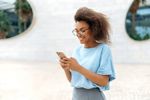 Gadget, wireless technology. Successful African American young curly woman with glasses, wearing blue shirt, standing outdoors near business center, using cell phone, texting, answering emails, smiles - Fotó, kép