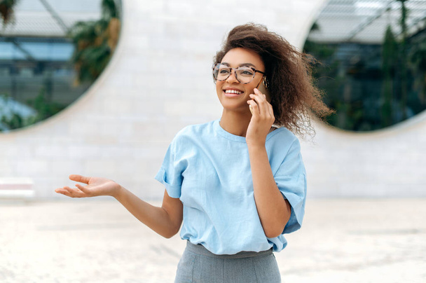 Happy young african american curly haired woman, wearing blue business shirt, with glasses, walking outdoors, going to a meeting, talking by smartphone, looking away, gesture hand, smiling friendly - Foto, immagini
