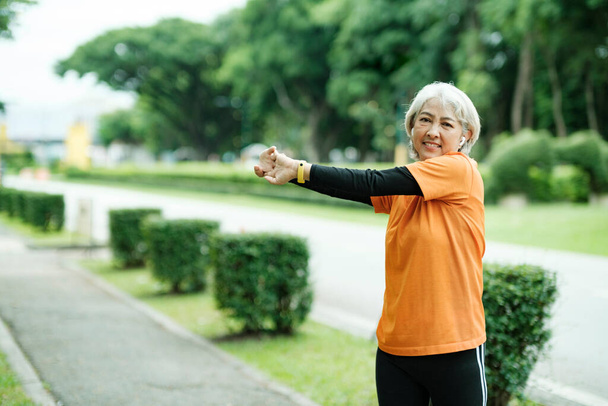 Athletic Senior woman jogger warming up by stretching arms and upper body before running in park. - Фото, изображение