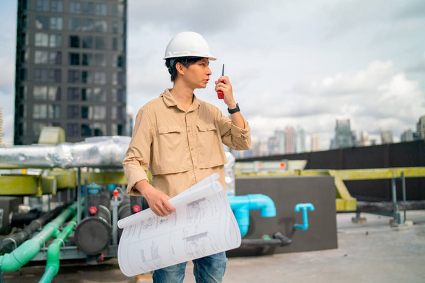 Ázsiai mérnök vagy technikus dolgozó gazdaság tervrajz használja walkie-talkie felvenni a kapcsolatot a munkatárs, hogy folytassa a munkát az építési területen, és ő marad az épület teraszán. - Fotó, kép