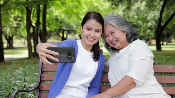 Пожилая мать и азиатская дочь отдыхают в парке Happy на отдыхе, держат смартфоны и фотографируются вместе. Здравоохранение для пожилых людей. старший по уходу - Кадры, видео