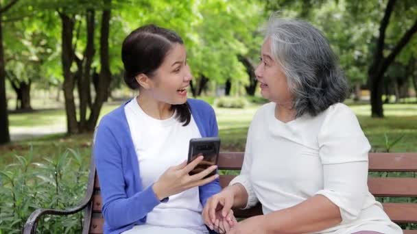 Пожилая мать и азиатская дочь отдыхают в парке Happy на отдыхе, держат смартфоны и фотографируются вместе. Здравоохранение для пожилых людей. старший по уходу - Кадры, видео