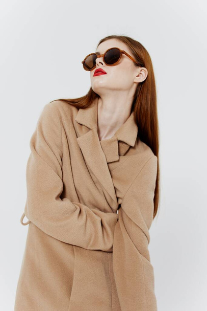 Una chica con estilo de pelo rojo impresionante y segura de sí misma en un abrigo y gafas posa en el estudio sobre un fondo blanco. Fotografía del catálogo publicitario de la revista. Foto de alta calidad - Foto, Imagen