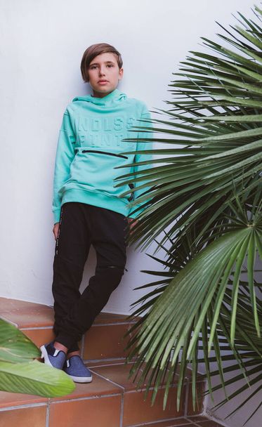 A teenager in sportswear stands on the steps against a white wall. Sports sweatshirt and black pants on the boy. Sports and fashion - Foto, afbeelding