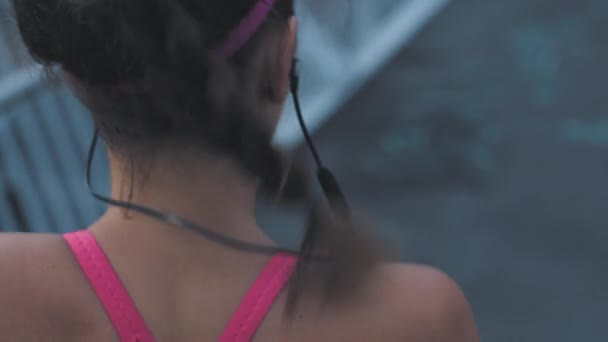 Fit woman running and jogging on a bridge in a city outdoors from behind. Closeup of a young athlete listening to music with earphones while doing cardio. Exercising for better health and fitness. - Metraje, vídeo