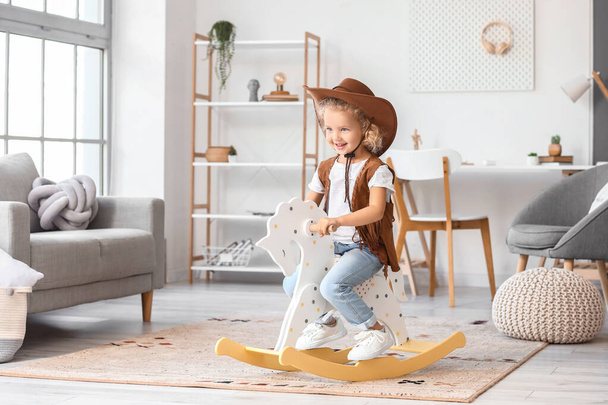 Cute little girl in hat and vest playing with rocking horse at home - Фото, зображення