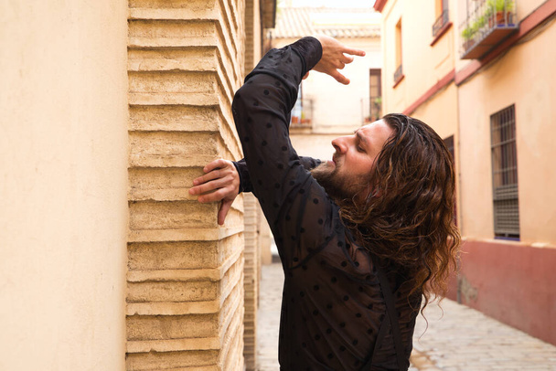 Homem de cabelos compridos dançando flamenco com camisa preta e rosas vermelhas. Ele faz posturas de dança com as mãos em uma típica rua estreita de Sevilha. Conceito de dança flamenca patrimônio cultural da humanidade. - Foto, Imagem