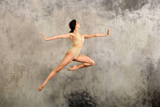 widok na całe ciało z boku kobieta baletnica w body skok do góry z rozkładanymi ramionami o szarą ścianę w studio - Zdjęcie, obraz