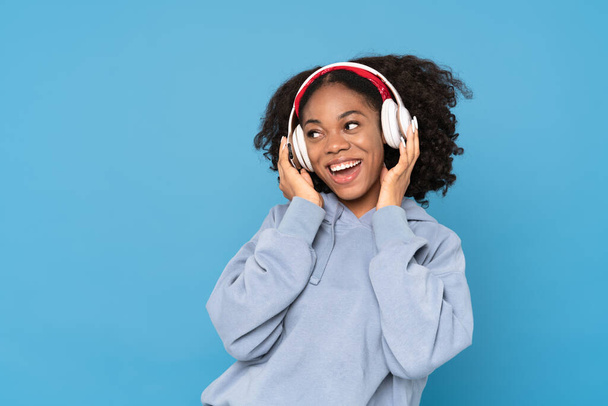 Mavi arka planda izole edilmiş kulaklıklarla müzik dinlerken gülümseyen genç siyahi kadın. - Fotoğraf, Görsel