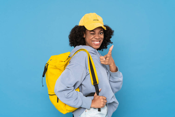 Black woman with backpack winking and pointing finger at camera isolated over blue background - Фото, изображение