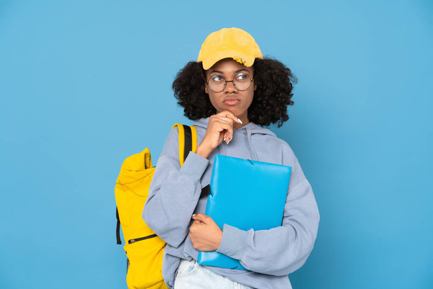 Black puzzlement woman posing with backpack and folder isolated over blue background - Photo, Image