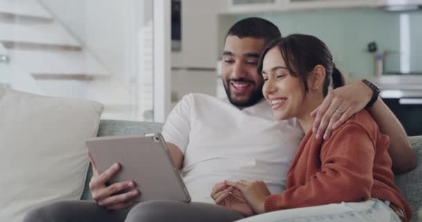 A couple talking on a tablet while sitting on a couch. A young inter racial man and woman video calling together indoors. People waving online - 映像、動画