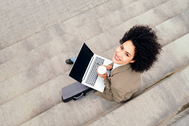 top view of a smiling woman working with laptop computer sitting outdoors on a staircase, concept of business and urban lifestyle, copy space for text - Valokuva, kuva
