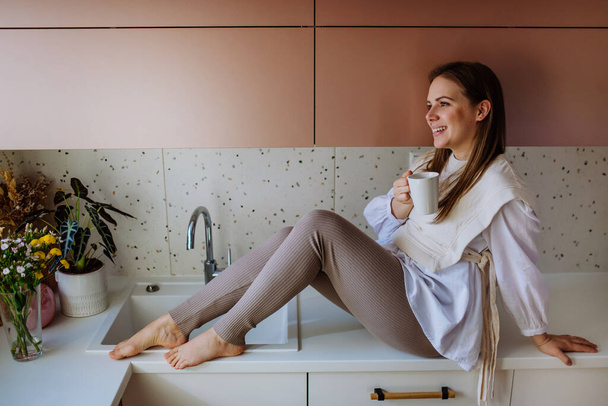A cheerful young woman holding coffee cup while sitting on the kitchen counter - Fotografie, Obrázek
