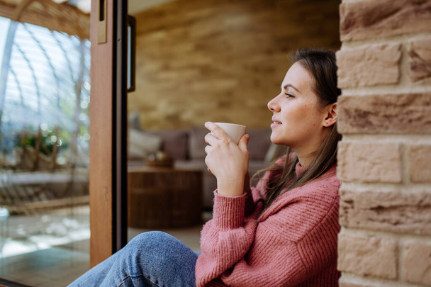 A smiling young woman with dreamy face drinking morning coffee and sitting at her home patio - Фото, зображення