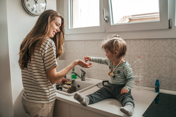 Little girl sitting on the kitchen sink helping her happy young mother to wash the cooking pot - Zdjęcie, obraz