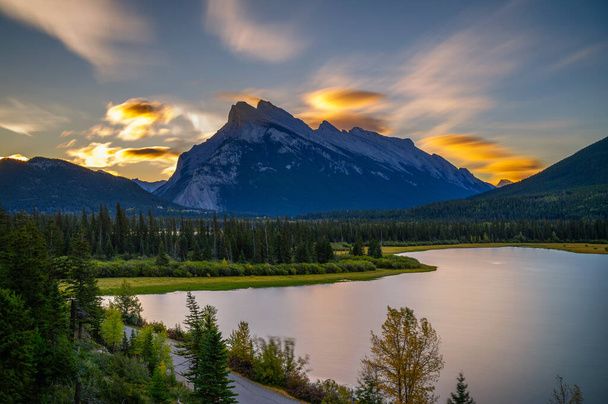 カナダのバンフ国立公園の朱色の湖の上にカラフルな日の出。謎の山が背景にある. - 写真・画像