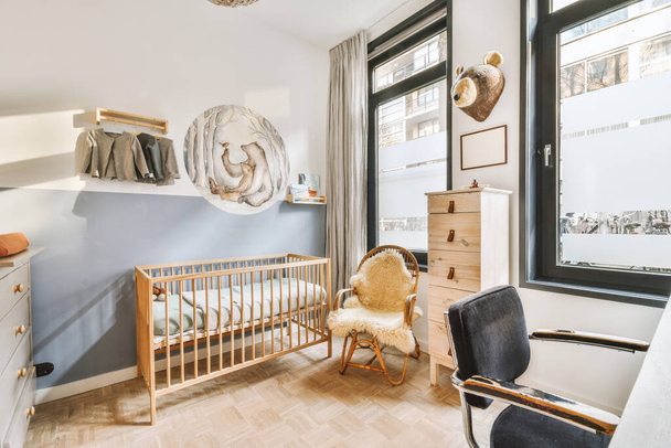 Интерьер белой комнаты с детской кроваткой и креслом возле окна с голубым ящиком при дневном свете - Фото, изображение