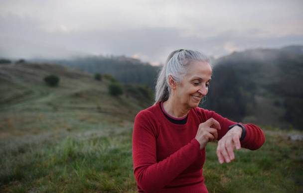 A senior woman jogger setting and looking at sports smartwatch, checking her performance in nature on early morning with fog and mountains in background. - Foto, Imagem
