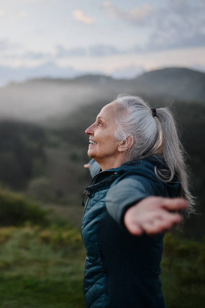 A senior woman doing breathing exercise in nature on early morning with fog and mountains in background. - Foto, Imagem