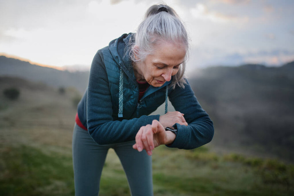 A senior woman jogger setting and looking at sports smartwatch, checking her performance in nature on early morning with fog and mountains in background. - Foto, imagen