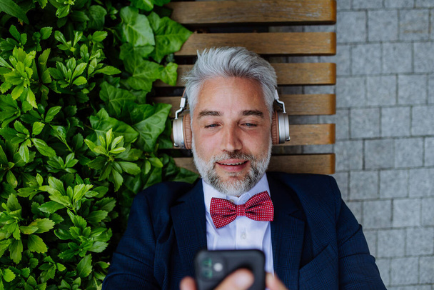 A mature businessman enjoying music in headphones relaxing on bench in city park during break at work. - Foto, afbeelding