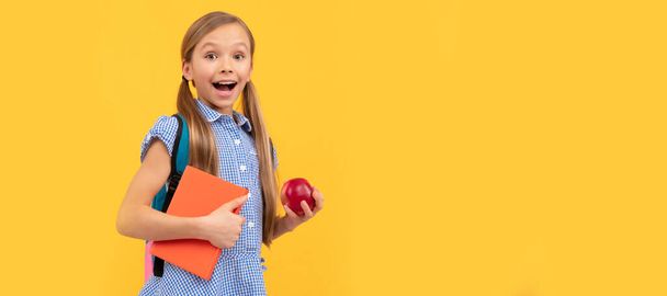 Yllättynyt tyttö lapsi pitää maukas omena terveen takaisin kouluun välipala, koulun hedelmiä. Koulutytön oppilaan lippu. Koululainen oppilas muotokuva kopio tilaa - Valokuva, kuva