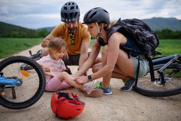 A mother and father helping their little daughter after falling off bicycle outdoors - Фото, изображение