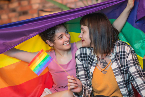 Very romantic lesbian couple on the sofa hugging each other and holding the LGBT rainbow flag on the back they spending amazing time together at home on the sofa. Portrait - Фото, зображення