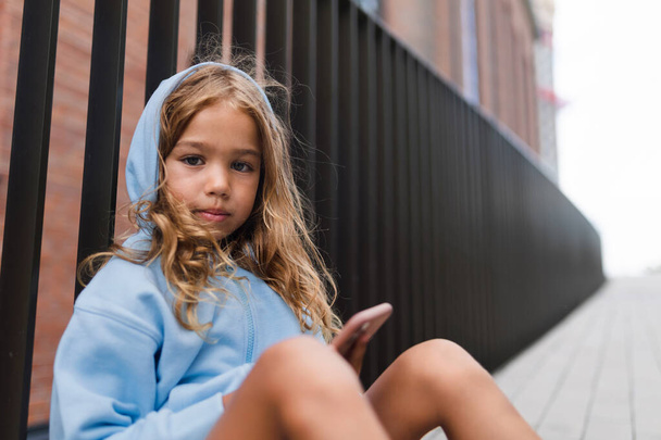 Little blond girl with hood sitting in sidewalk, holding a smartphone, looking at camera. - Φωτογραφία, εικόνα