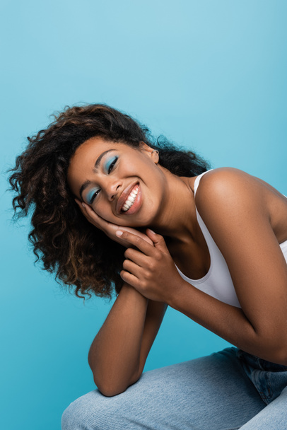 cheerful african american woman with blue eye shadows holding hand near face and looking at camera isolated on blue - Фото, зображення